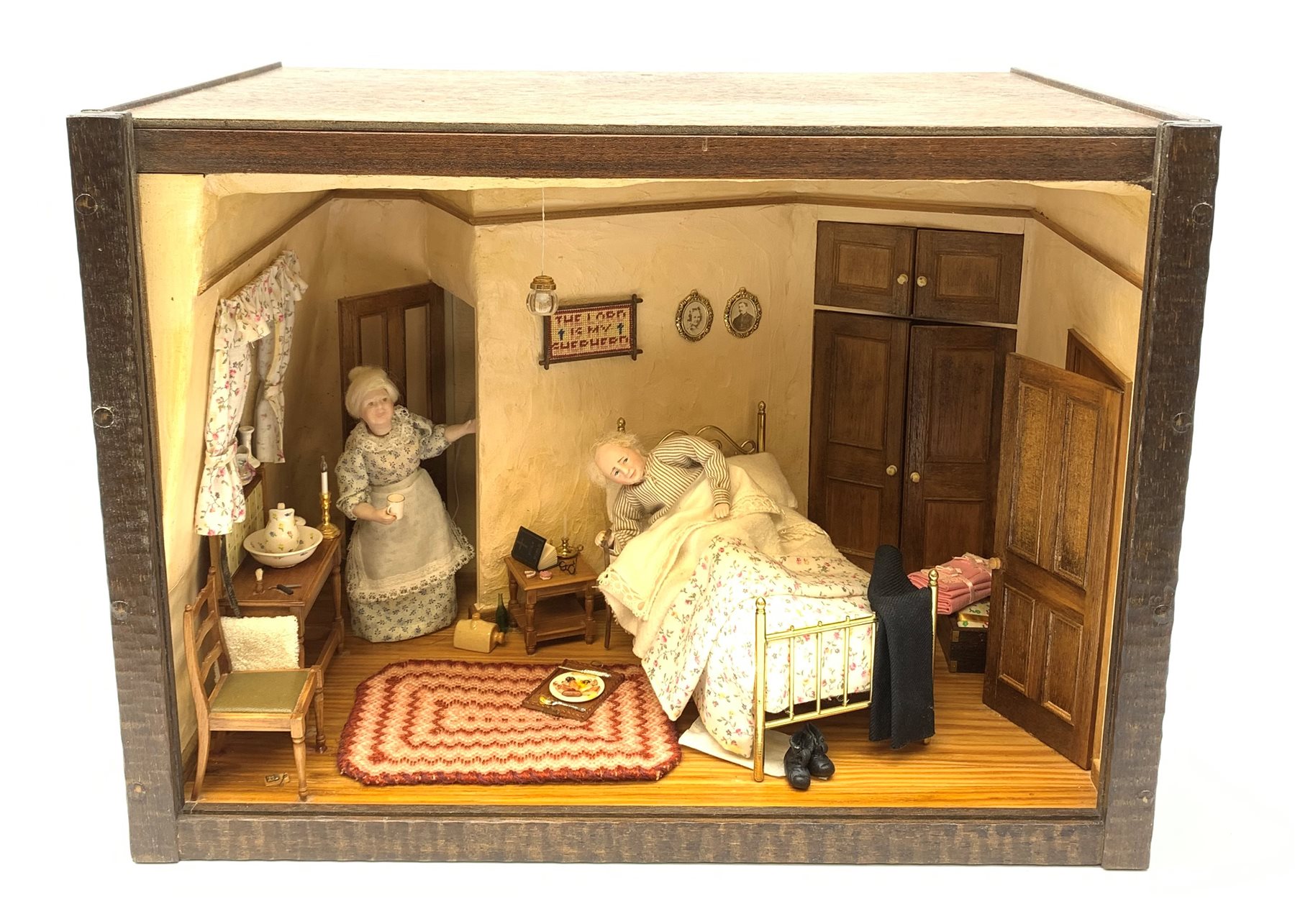 A 1/12th scale diorama of a cottage bedroom, depicting an elderly couple, he in bed, her by the door with cup in hand, the interior furnished with wash stand, wash jug and bowl, needle work, brass bed, and mouse trap under chair, plus light up candle and ceiling pendants, etc., H29cm L40cm D26cm.


