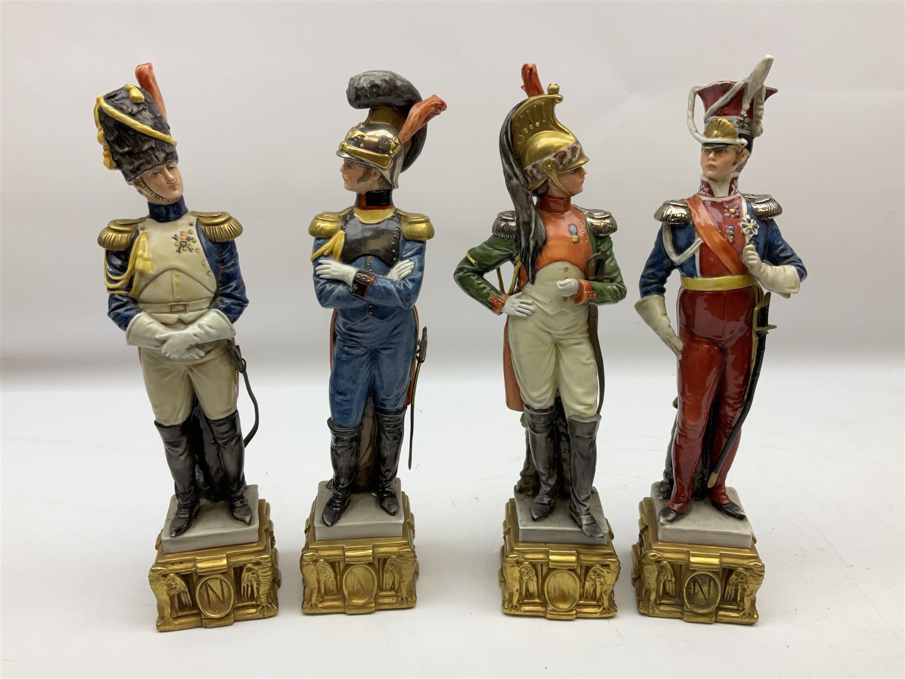 Group of seven Capodimonte figures of soldiers, to include four Bruno-Merli examples including Officer of the 12th Lancers 1820 and Captain of the 18th Hussars 1815, both raised upon ornate gilt plinths bearing British monarch motto in Latin 'Dieu et mon droit', and three further similar, all with crowned N mark beneath, tallest H32cm