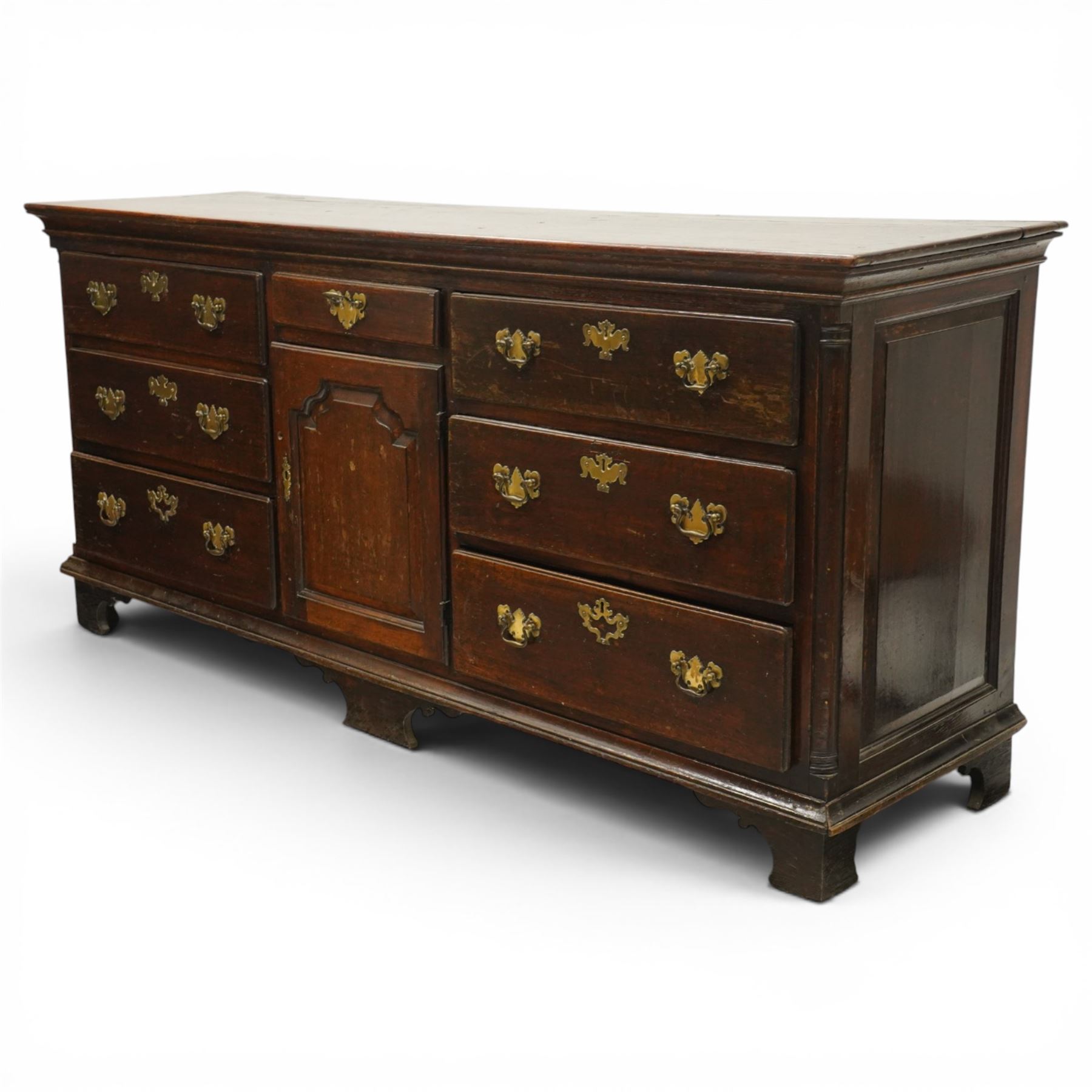 19th century Georgian oak long dresser base, rectangular plank top above a central frieze drawer over a fielded panel cupboard, flanked by three banks with quarter cantered pilasters, brass swan neck handles and escutcheons, raised on shaped bracket feet