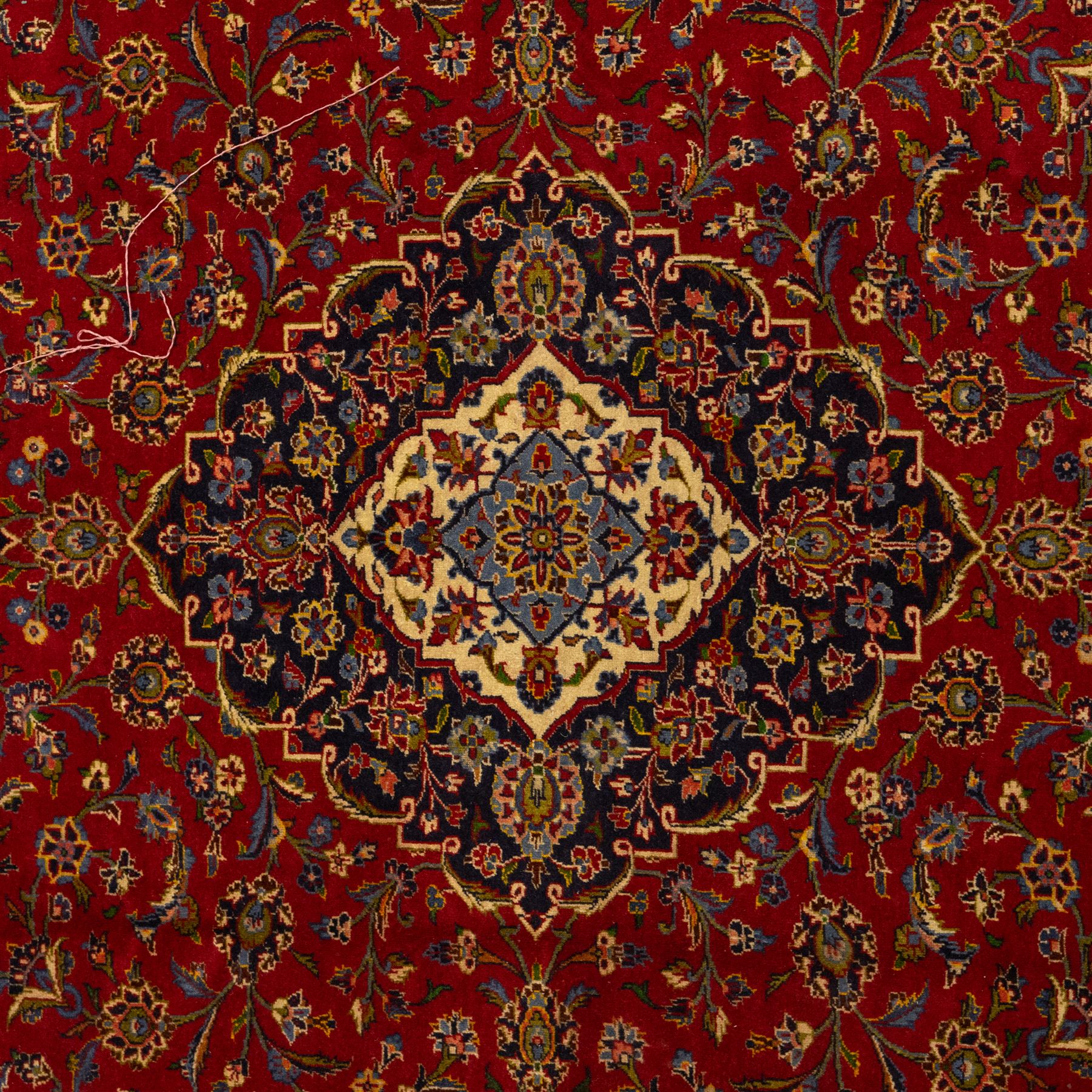 Central Persian Kashan hand-knotted carpet, crimson ground, with a central shaped palmette medallion with pendants, surrounded by scrolling foliate and floral motifs incorporating rosettes, palmettes, and vine work, enclosed by a dark navy with repeating border with repeating palmettes and meandering vines, flanked by floral guard stripes, with cotton fringes 