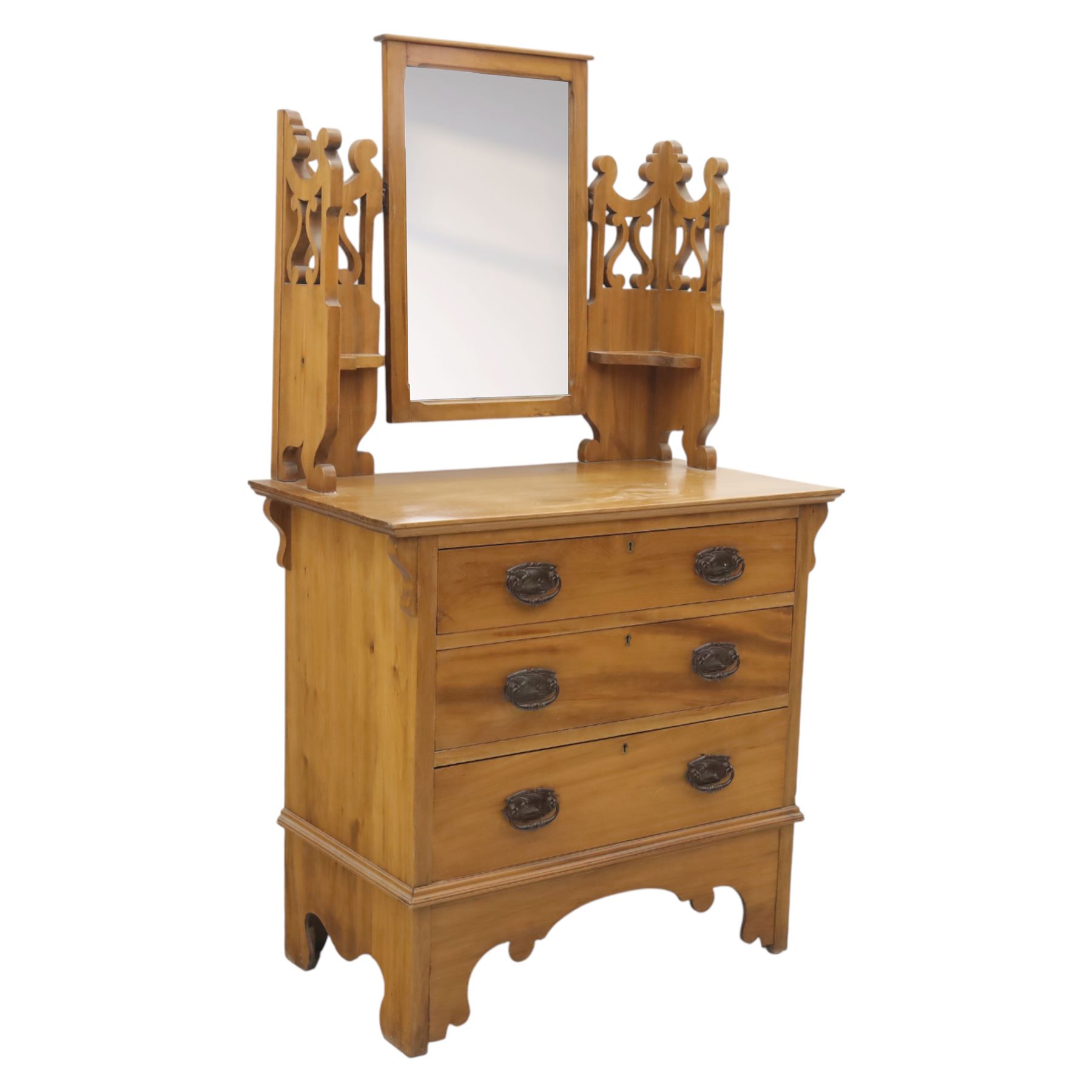 Edwardian stained pine dressing chest, the moulded top over three graduating drawers with pressed brass handles, shaped apron and bracket feet, the mirror back with rectangular plate on pivot supports, flanked by pierced Gothic style uprights with small corner shelves