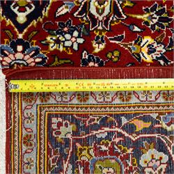 Central Persian Kashan hand-knotted carpet, crimson ground, with a central ivory shaped floral medallion with pendants, surrounded by dense scrolling foliate and floral motifs incorporating rosettes, palmettes and delicate vine work, enclosed by ivory spandrels and a dark navy border with repeating palmettes and meandering vines, flanked by floral guard stripes, with cotton fringes