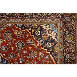 Central Persian Kashan hand-knotted rug, madder red field with central articulated cusped medallion in ivory and sky blue, issuing network of scrolling arabesques palmettes and floral sprays with conforming spandrels, indigo main border of scrolling vinery and stylized palmettes, framed by multiple guard borders, with cotton fringe ends  