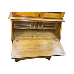 Early 20th century Arts and Crafts oak bureau bookcase, twin leaded and glazed stained glass doors enclosing two shelves, over bureau section with fall-front inlaid with sunburst motif, fitted interior of pigeonholes and shelves, on bracket feet