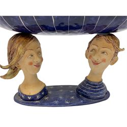 Ceramic 'girl and boy dish', by Helen Kemp, an oval dish with a mottled pink ground and gilt edging upon the sculpted heads of a girl and a boy, together with a 'boy sconce', by Helen Kemp, a sculpted boy holding a small bowl with foliate decoration, both with the artist's mark beneath.