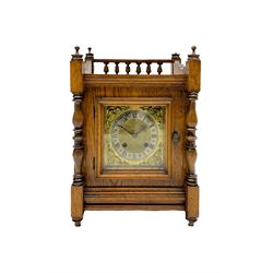 An early 20th century oak cased mantle clock in the arts and crafts style, case decorated with a rear balustrade, turned finials and flanking ring turned pilasters to the dial, square brass dial with cherub spandrels, silvered chapter ring, matted dial centre and gothic steel hands, with a German eight-day spring driven countwheel movement striking the hours and half hours on a coiled gong. With pendulum. 

