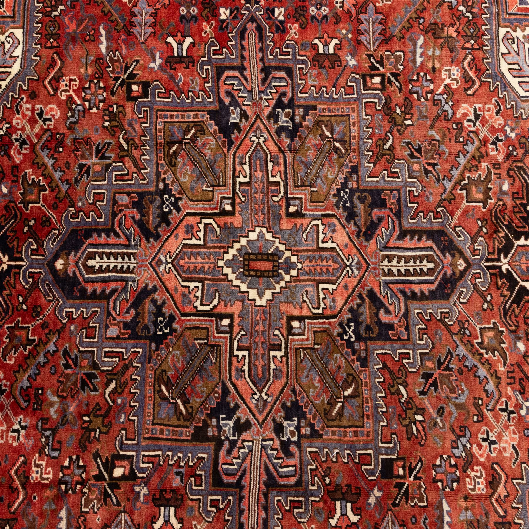 North West Persian Heriz hand-knotted carpet, madder red field with central stepped geometric medallion with radiating angular motifs and serrated outlines, and stylized floral and tribal devices framed by a broad border of repeating angular palmettes and rosettes flanked by multiple guard stripes, with cotton fringe ends 