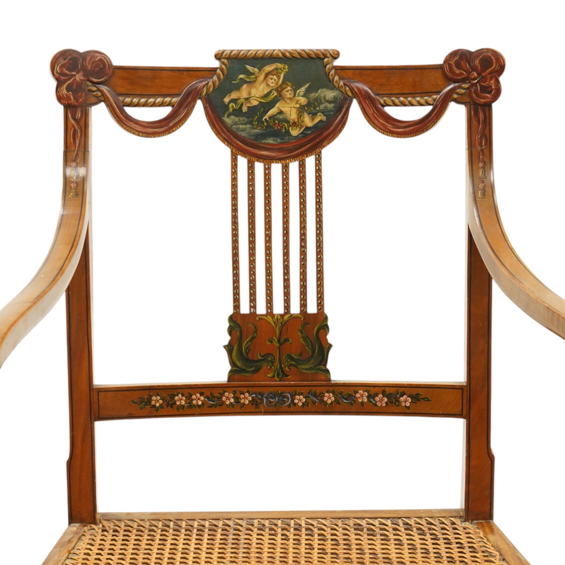 Late 19th century Adam Revival painted and parcel-ebonised satinwood open armchair, the cresting rail centred by a painted panel depicting putti holding garlands, within draped swag ornament and moulded ribbon-twists, over a pierced vertical splat with gilt beaded detail, the downswept arms and caned seat above a painted lattice and floral frieze, raised on ring-turned tapering front supports with musical instrument painted panel and acanthus leaves
