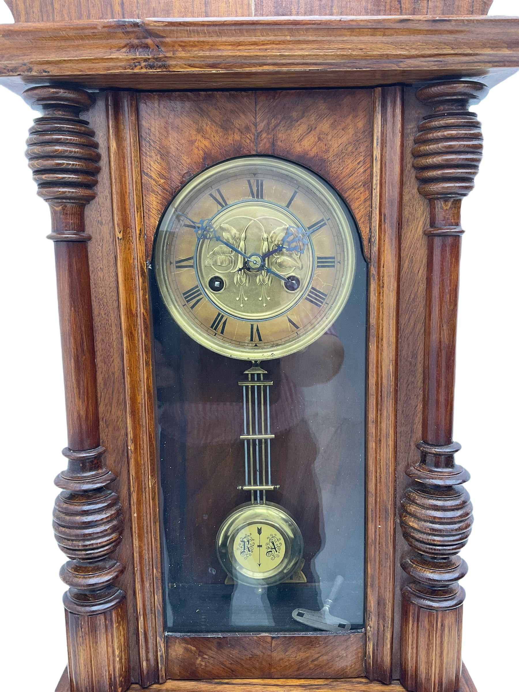 German early 20th century wall clock in a mahogany case with an architectural pediment and turned finial, with applied carving and pendant finials, fully glazed door with a visible gridiron pendulum, eight-day spring driven striking movement striking the hours on a gong, dial with Roman numerals to the chapter and a gilt repoussé centre.   

