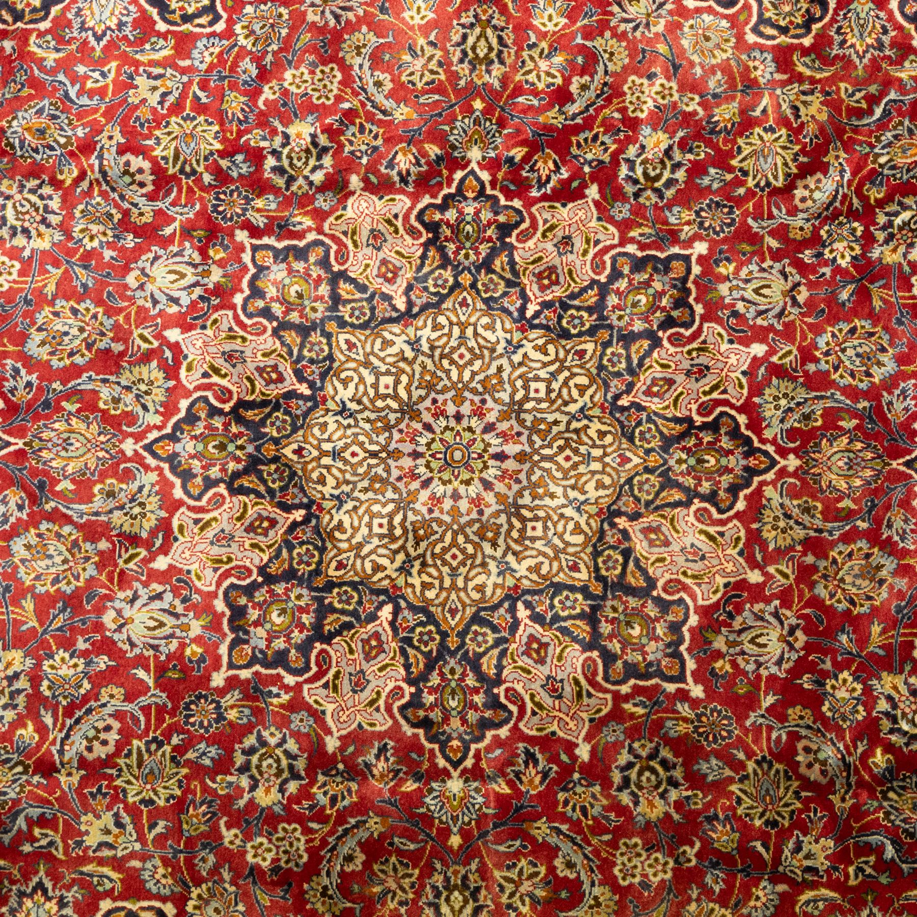 Central Persian Kashan hand-knotted carpet, crimson ground, with a large central ivory and dark navy floral medallion with pendants, surrounded by dense scrolling foliate and floral motifs incorporating rosettes, palmettes and vine work, enclosed by ivory spandrels and a dark navy border with repeating palmettes and meandering vines, flanked by floral guard stripes