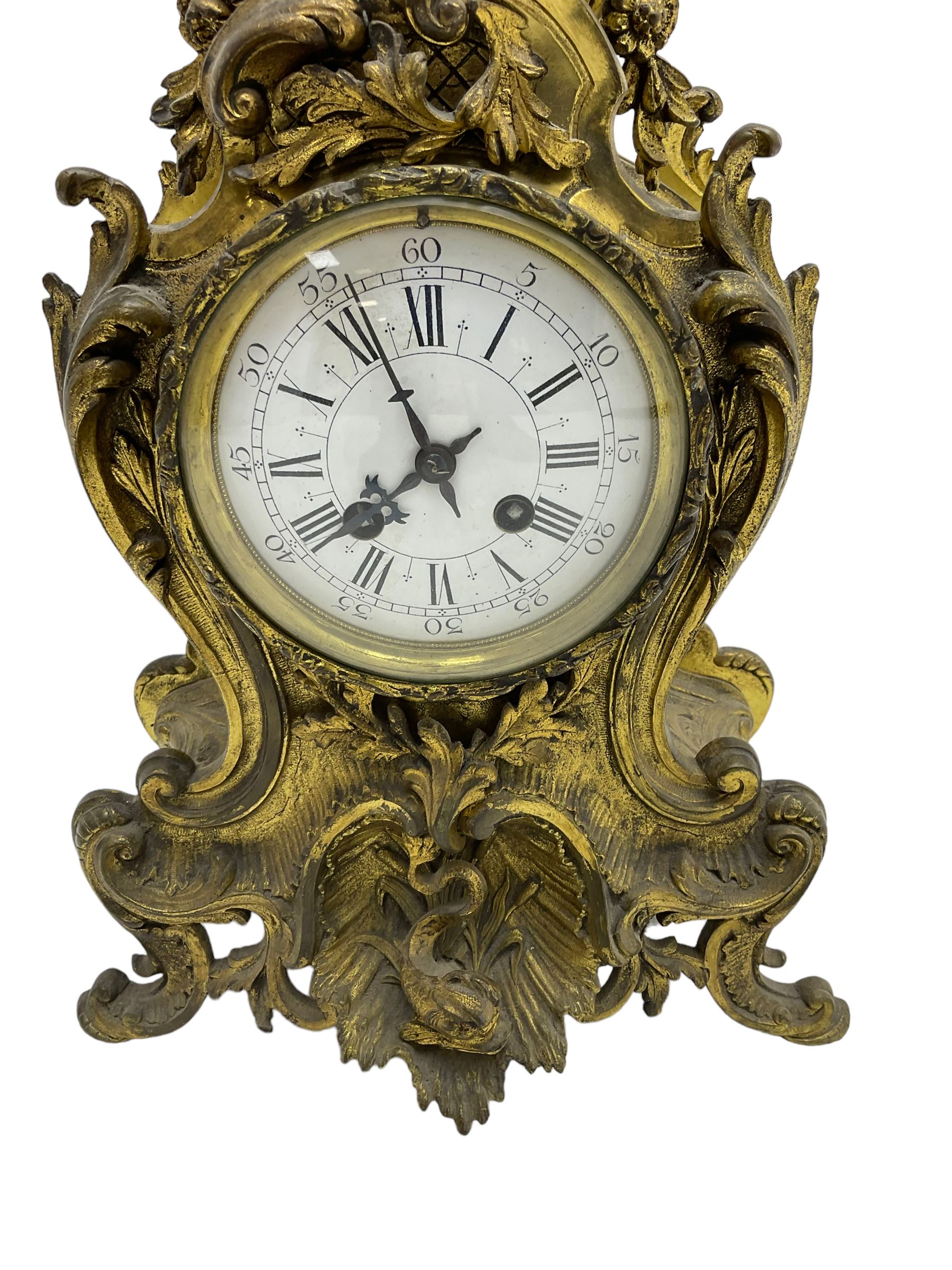 French - late 19th century 8-day gilt brass mantle clock surmounted by an urn, with a waisted case and baroque decoration raised on four splayed feet, with a glazed bezel, enamel dial with Roman numerals, five minute Arabic's and gothic steel hands, count wheel striking movement, striking the hours and half-hours on a bell. With pendulum.