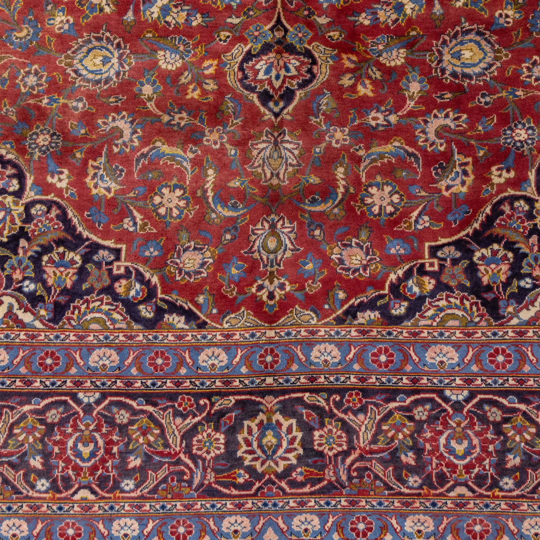 Central Persian Kashan hand-knotted carpet, madder red field with a central cusped medallion of ivory and deep indigo ground, enclosing a stylized floral rosette, framed by scrolling arabesques, field richly ornamented with an intricate network of flowering vinery, palmettes, and delicate floral sprays, spandrels in deep indigo, principal border of navy ground decorated with a continuous palmette and vine-scroll, flanked by multiple guard stripes, with cotton fringe ends 