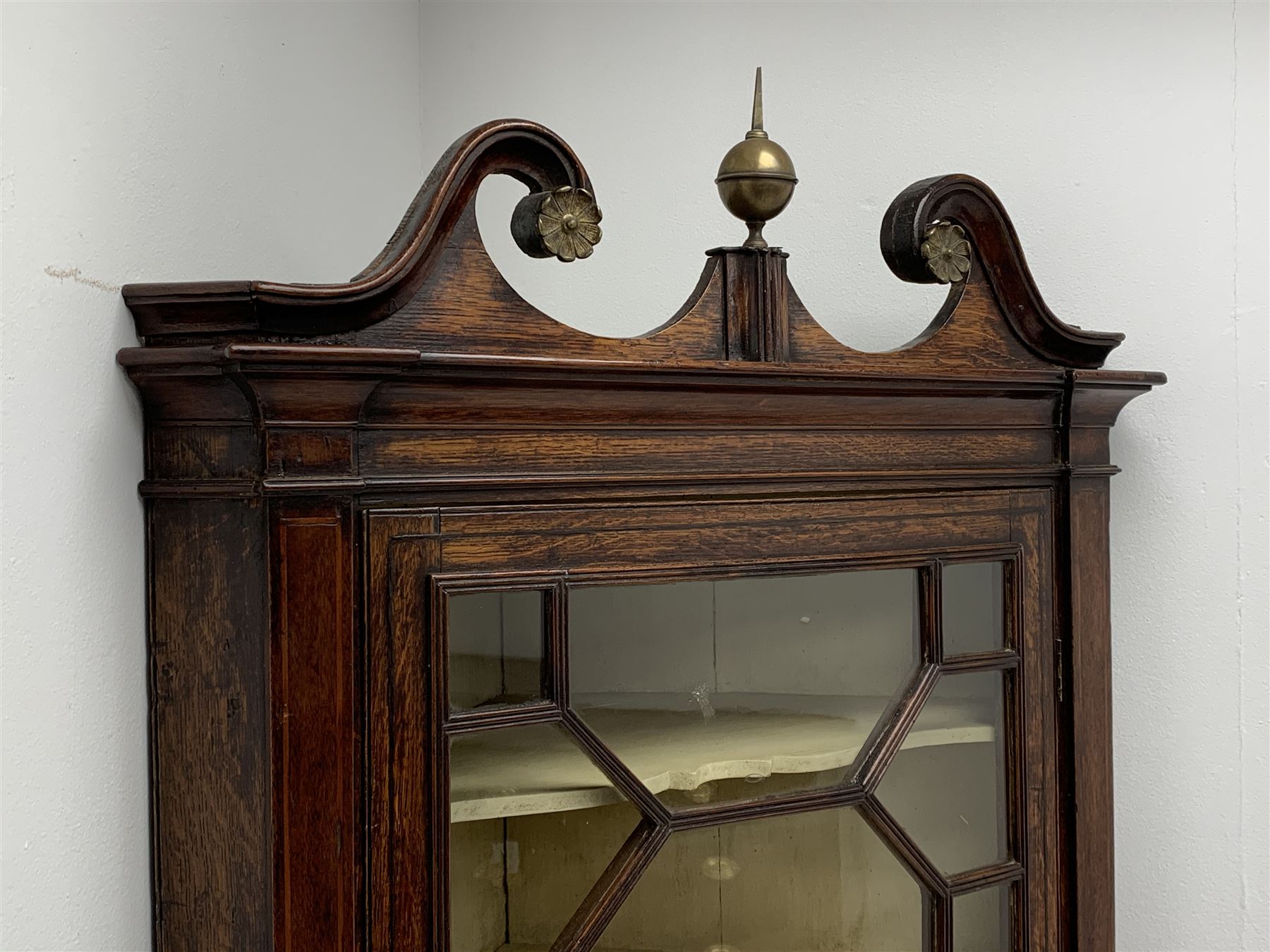 George III oak floor standing corner cabinet, scrolled swan neck pediment with central finial over astragal glazed door, painted interior fitted with three shaped shelves, the lower section fitted with single shaped shelf enclosed by two fielded panel doors inlaid lozenges, on bracket feet, H195cm, W77cm