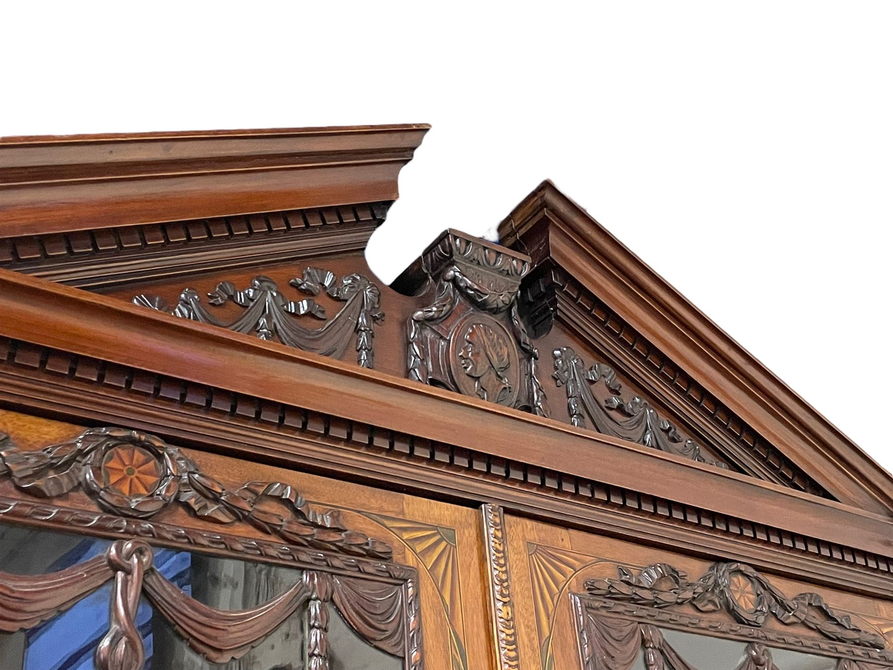 Large and impressive late 19th century Irish mahogany secretaire bookcase, moulded sloped arch dentil pediment decorated with central carved mask surrounded by linen festoons and trailing bellflowers, canted and fluted uprights with carved acanthus leaf upper and lower brackets, enclosed by two astragal glazed doors in the form of trophies with twist ribbon carved mouldings, inlaid with fanned spandrels and trailing leaf painted satinwood band, cylinder roll top inlaid with central fan motif and fanned spandrels, pull-out action revealing stepped fitted interior, central tambour roll cupboard flanked by two Composite pilasters and a combination of drawers and pigeon holes, fitted with three break-front drawers, matched canted and fluted corners with acanthus leaf carvings, on square tapering and fluted spade feet