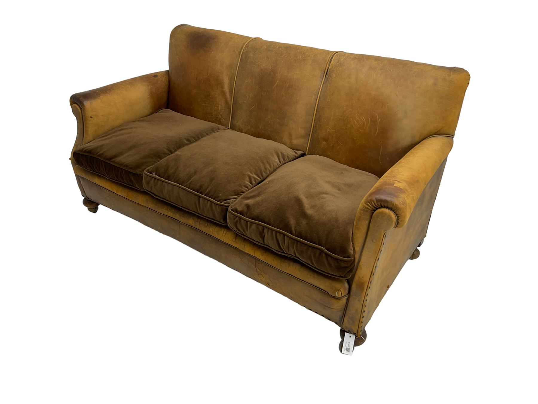 Early-to-mid-20th century three seat sofa, upholstered in worn tan leather with studwork, loose cushions upholstered in brown fabric, on compressed bun feet with castors