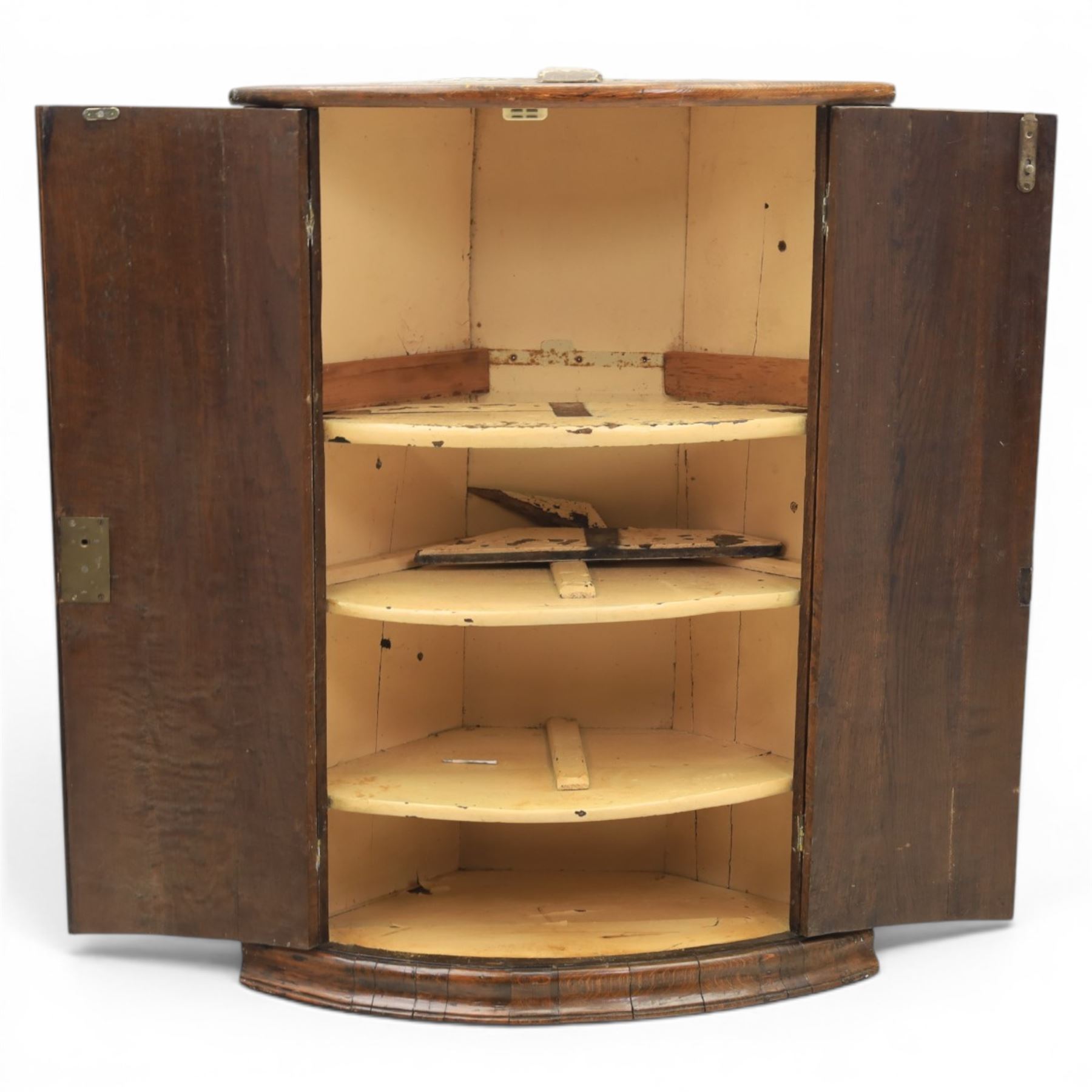 George III oak cylindrical corner cupboard, bow fronted doors revealing a painted shelved interior, brass escutcheons  and hinges, raised on a moulded plinth base