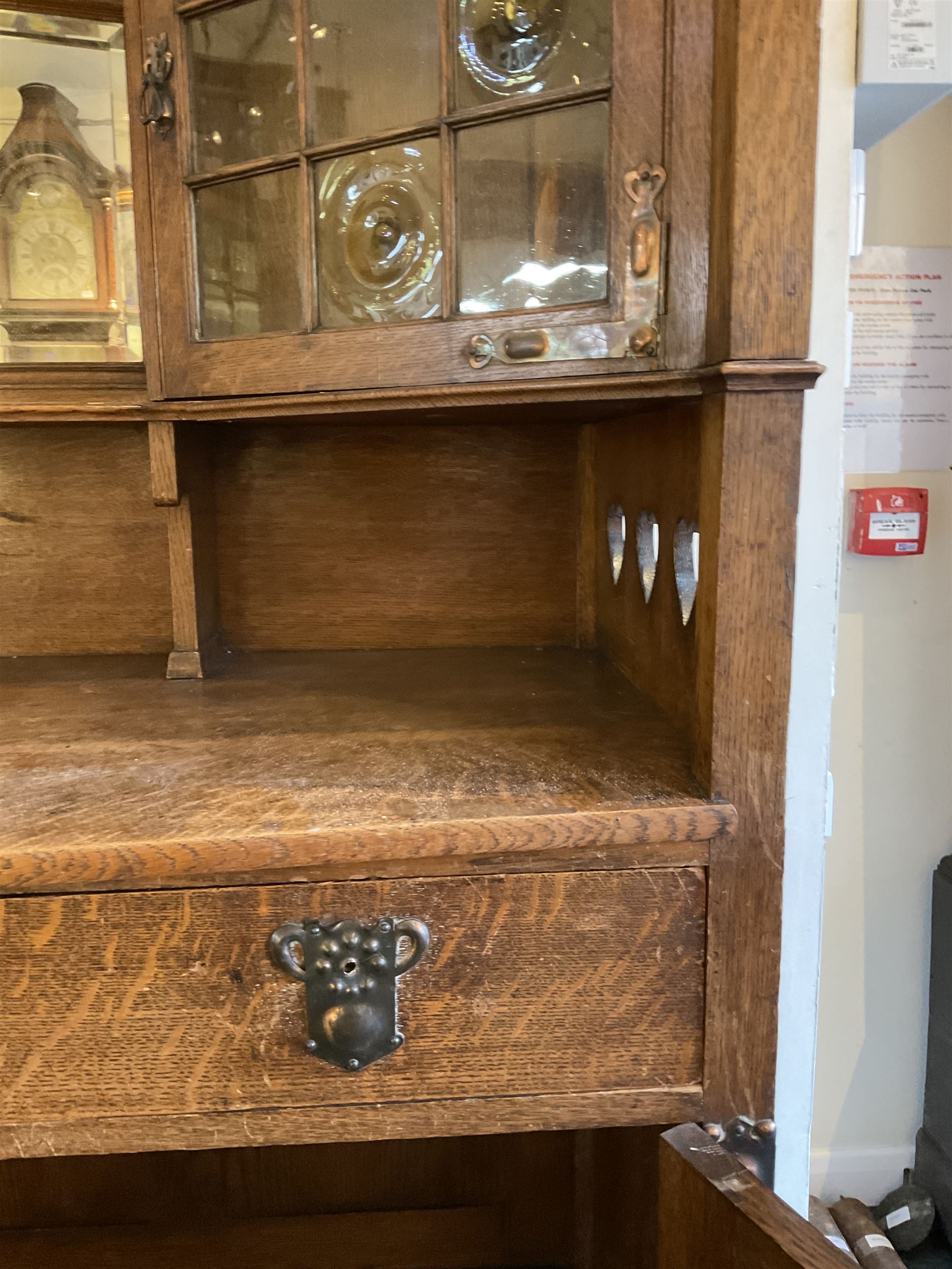 Shapland and Petter - Arts & Crafts period oak dresser, raised gallery top with spade balustrade, central bevelled mirror flanked by two cabinet, astragal glazed doors with yellow tinted glass and bullseye panes, the sides with pierced heart shaped decoration, fitted with two drawers and two double cupboards, enclosed by linenfold panelled doors,  copper work strap hinges and fixtures, on shaped spade feet, the locks stamped 'S&P'