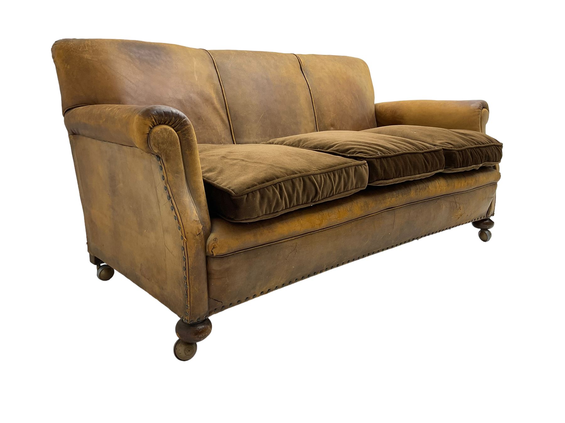 Early-to-mid-20th century three seat sofa, upholstered in worn tan leather with studwork, loose cushions upholstered in brown fabric, on compressed bun feet with castors