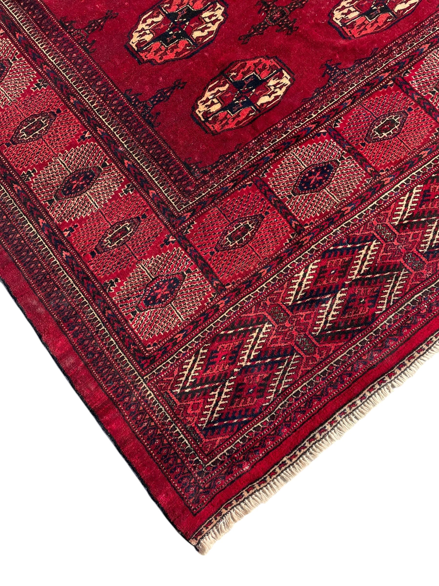 Afghan Bokhara red ground carpet, the field decorated with four rows of Gul motifs interspersed with Hac motifs, the border with lattice pattern panels within small geometric guard stripes, geometric patterned end panels