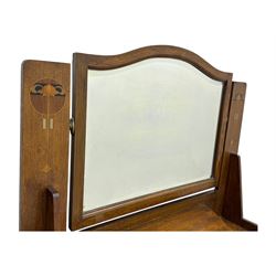 Early 20th century Arts and Crafts oak dressing chest, the arched mirror flanked by floral marquetry inlaid uprights above a shelf and drawer, over three long drawers with square hammered metal plates and circular pulls, on slender supports with castors