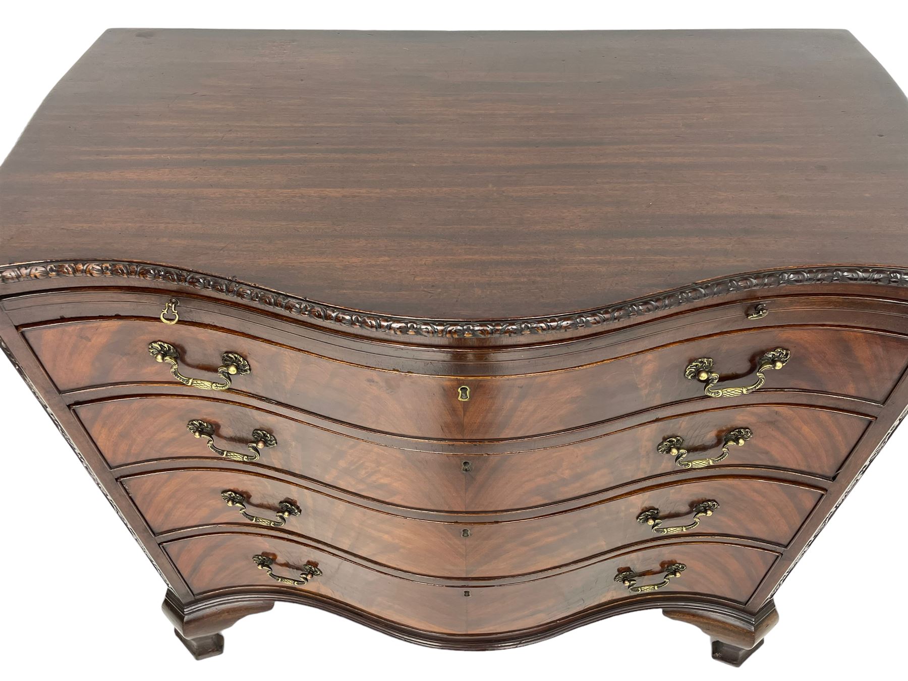 Chippendale design serpentine chest, shaped moulded top carved with repeating flower head motifs, fitted with brushing slide over four graduating drawers, pierced and foliate moulded handle plates with scrolled swan neck handles, canted upright corners carved with flower heads and strapwork, on canted ogee bracket feet
