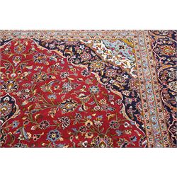Central Persian Kashan hand-knotted carpet, crimson ground field, with central cusped lozenge medallion, enclosing a palmette and a floral rosette arrangement, surrounding field densely populated with scrolling arabesques, flowering vinery and stylized boteh motifs in tones of indigo, pale blue, ivory and ochre and spandrels of confirming design, flanked by multiple guard stripes, with cotton fringe ends 