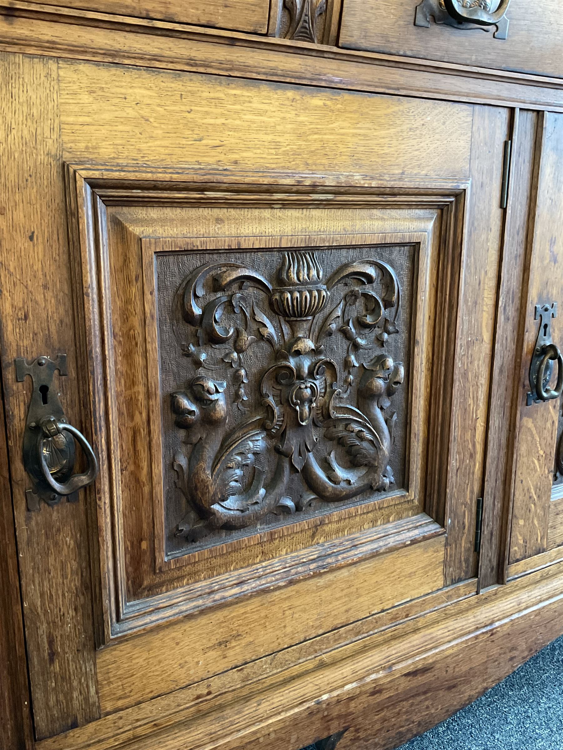 Late 19th century Arts & Crafts period oak mirror back sideboard, shaped bevelled mirror back carved with foliate scrolls and flower heads, down swept supports with elongated club-shaped uprights, moulded rectangular top over two drawers and three cupboards, the drawer fronts with copper handles decorated with sailing ships, panelled doors carved with mythical beasts, flowerheads and foliage, on moulded block feet