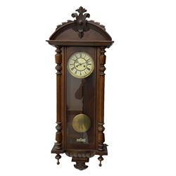 A late 19th century German Wall clock in a mahogany case with a carved semi-circular pediment, fully glazed door with turned side columns and pendant finials, two-part enamel dial with Roman numerals and steel gothic hands, eight-day spring driven movement sounding the hours on a coiled gong, with a wooden pendulum rod and spun brass bob, With key.

