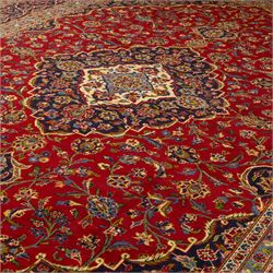 Central Persian Kashan hand-knotted carpet, crimson ground, with a central shaped palmette medallion with pendants, surrounded by scrolling foliate and floral motifs incorporating rosettes, palmettes, and vine work, enclosed by a dark navy with repeating border with repeating palmettes and meandering vines, flanked by floral guard stripes, with cotton fringes 
