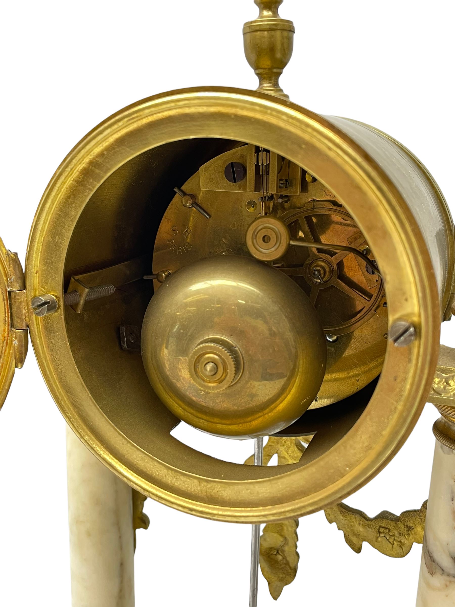 20th century French portico clock c1910 on a white veined marble base, gilt drum movement case supported between two marble columns with brass capitals and a suspended garland swag with a starburst pendulum beneath, enamel dial with Arabic numerals, floral decoration and Louis XV pierced gilt hands, convex glass and brass bezel, 8-day countwheel striking movement striking the hours and half hours on a bell. 




