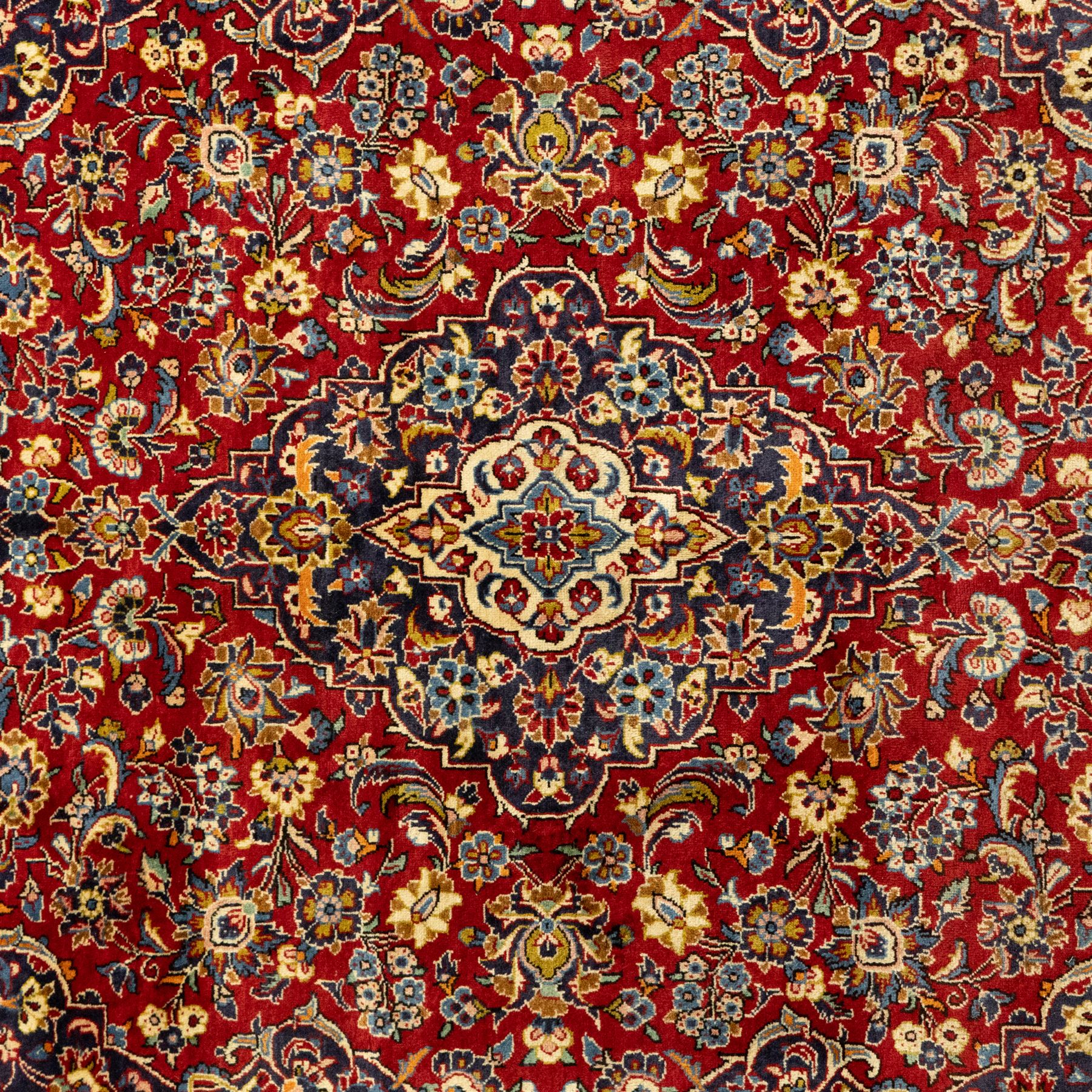 Central Persian Kashan hand-knotted carpet, crimson red central field with central floral medallion with floral vine-work and palmettes, within dense floral spandrels and a wide navy blue border decorated with continuous foliate motifs and multiple guard borders, with cotton fringe ends  