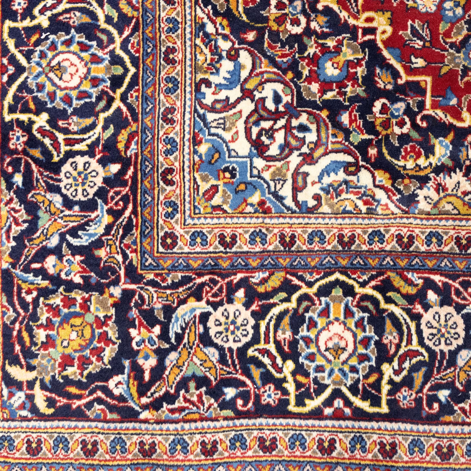Central Persian Kashan hand-knotted rug, rich red field with a central lobbed medallion surrounded by finely drawn scrolling floral sprays, palmettes, and rosettes, enclosed by a broad dark blue floral border with multiple guard stripes and cotton fringe ends 