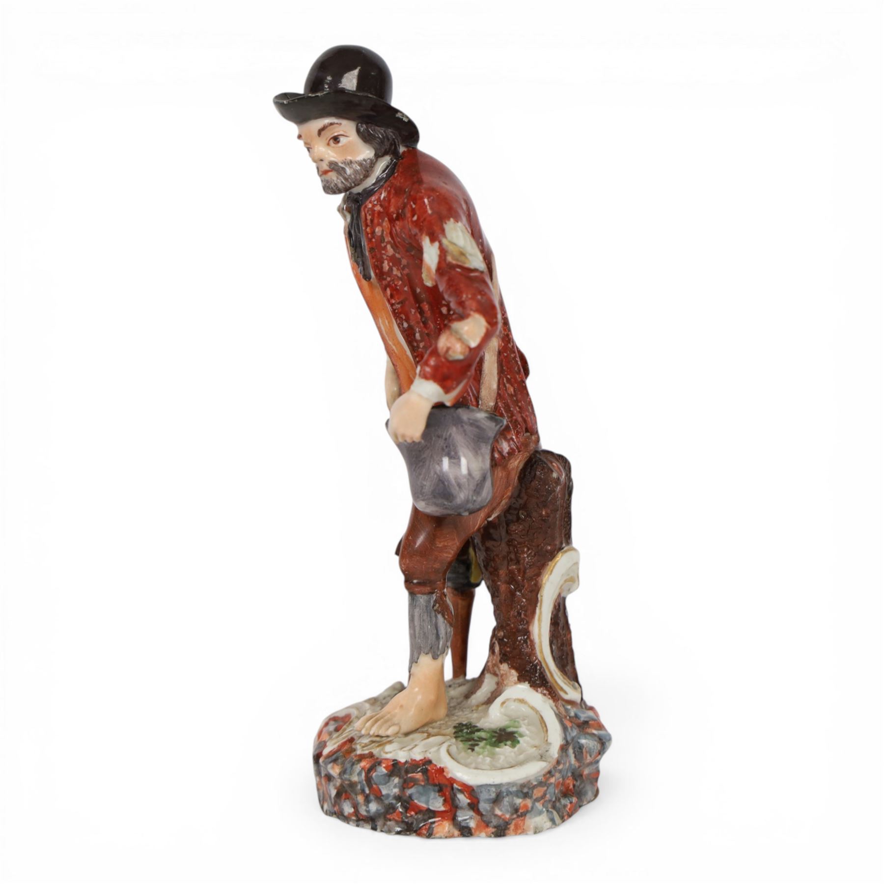 Mid 18th century Furstenberg figure, modelled as a beggar or mendicant with peg leg and bare foot, circa 1755, modelled by Simon Feilner, wearing tattered clothes, one arm supporting an alms pouch, the other holding a crutch, upon a part scrolled base, bearing paper labels beneath including one inscribed 'Sammlung Wilhelm Gumprecht 322', H15cm
