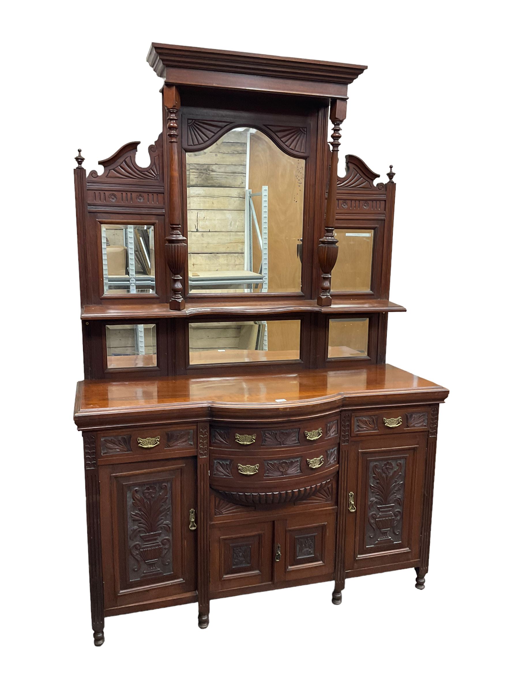 Edwardian walnut sideboard, projecting cornice supported by turned supports, the back fitted with three bevelled mirrors beneath fluted and flowerhead carved friezes, the lower section with four drawers and four panelled cupboard doors carved with foliate and urn designs, fluted uprights on turned feet