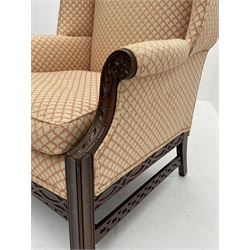 Georgian style mahogany framed wingback armchair, the scrolled arm terminals and supports carved with flower heads and foliage, sprung seat and back upholstered in pale lozenge patterned fabric, loose seat cushion, blind fretwork frieze rails, square moulded supports joined by pierced stretcher rails 