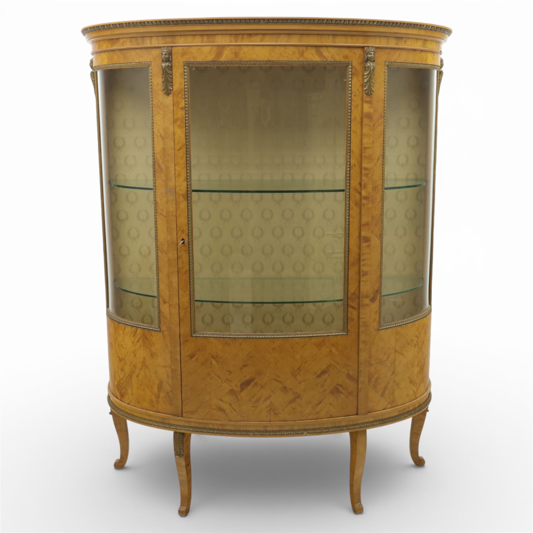 Edwardian satin walnut display cabinet, bowed oval form with moulded and dentil cornice over glazed panels, enclosing a laurel wreath damask interior with adjustable glass shelves, the frame applied with gilt metal bead mouldings and mask mounts, enclosing a shelved interior, raised on cabriole supports with moulded feet