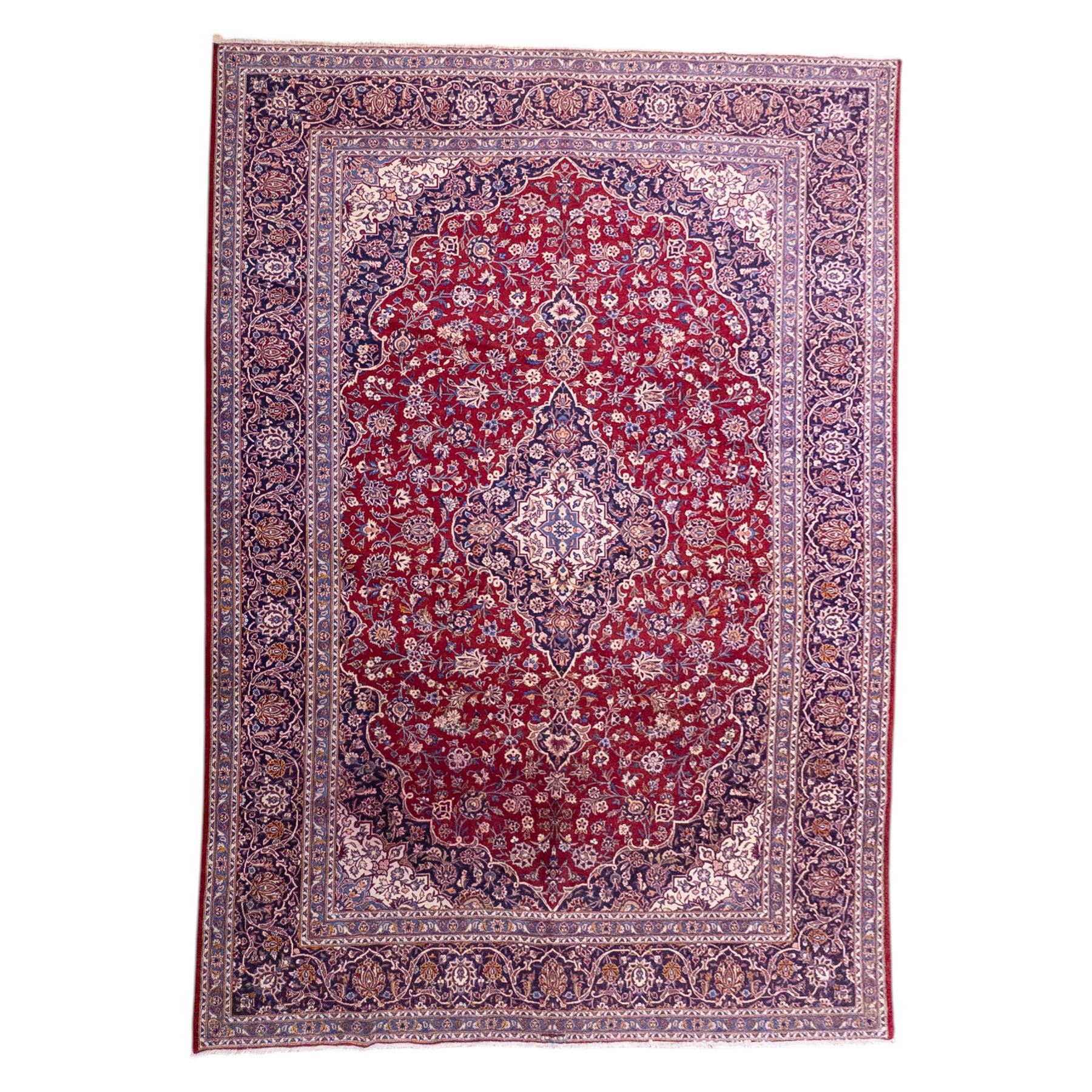 Central Persian Kashan hand-knotted carpet, madder red field with central cusped medallion in navy and ivory enclosing a floral rosette and radiating scrolling arabesques and palmettes, across a densely ornamented ground of finely drawn vinery enriched with a profusion of flowering sprays and stylized blossoms, spandrels in deep indigo and principal border of navy ground decorated with a continuous palmette and vine-scroll flanked by multiple guard stripes, with cotton fringe ends