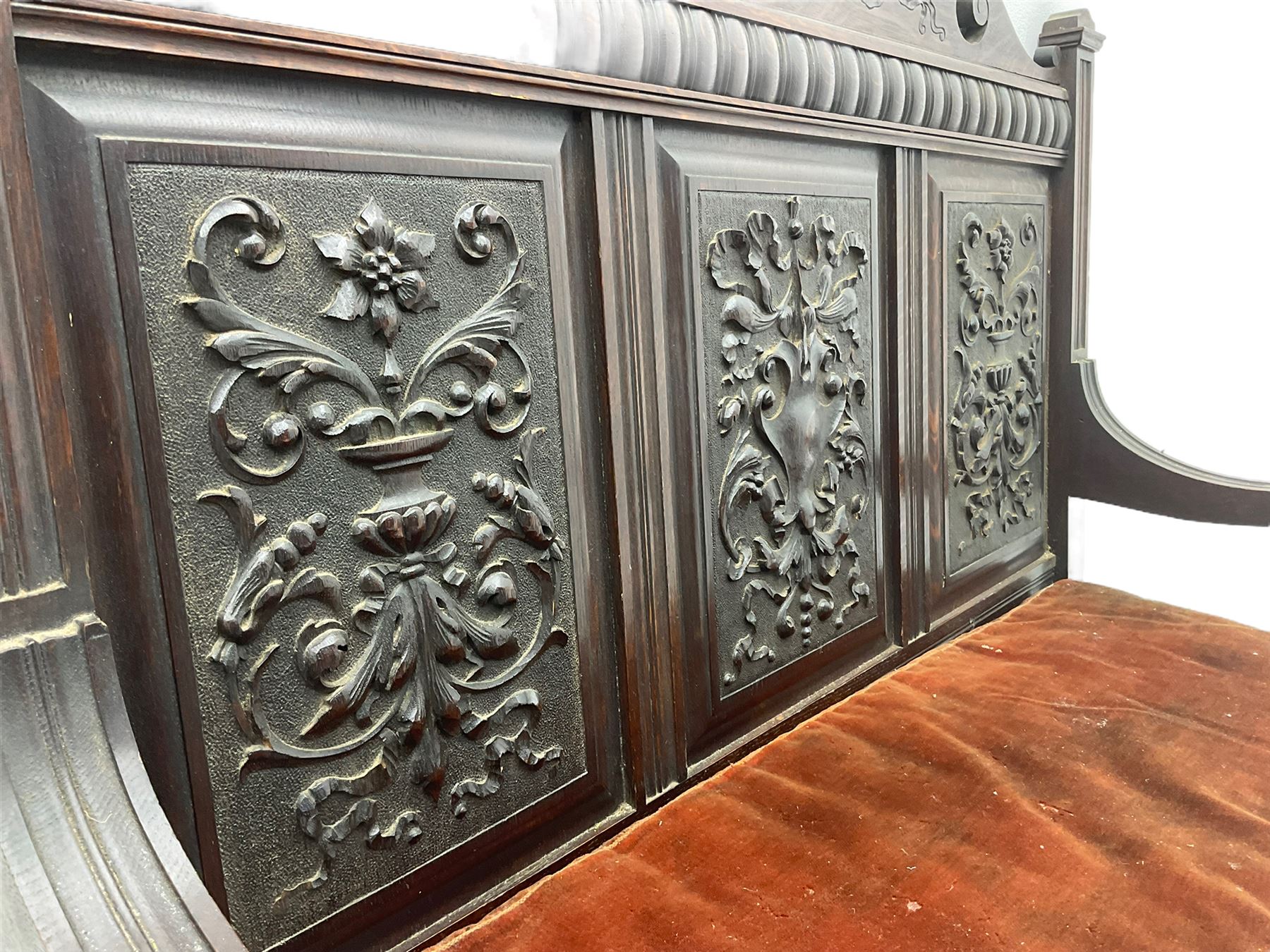 Late 19th century oak settle bench, shaped cresting rail carved with ribbon over triple panelled back, the panels relief carved with scrolled and floral urns and cartouche, hinged box seat, turned front feet, with upholstered squab cushion