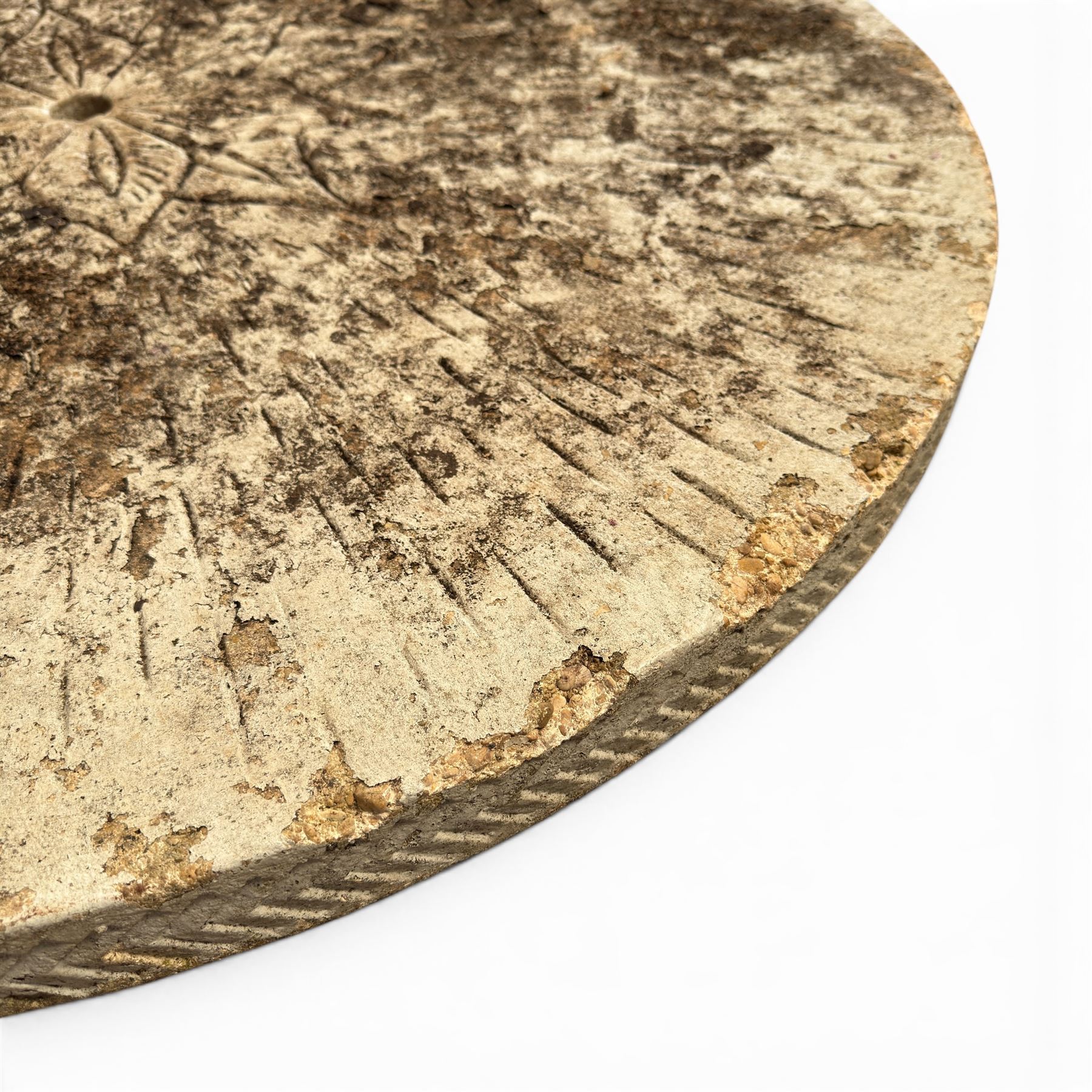 Weathered cast stone garden table, circular top etched with central floral medallion, the edge with repeating chevron motifs, raised on a quadriform S-scroll pedestal with stepped and chevron carved base