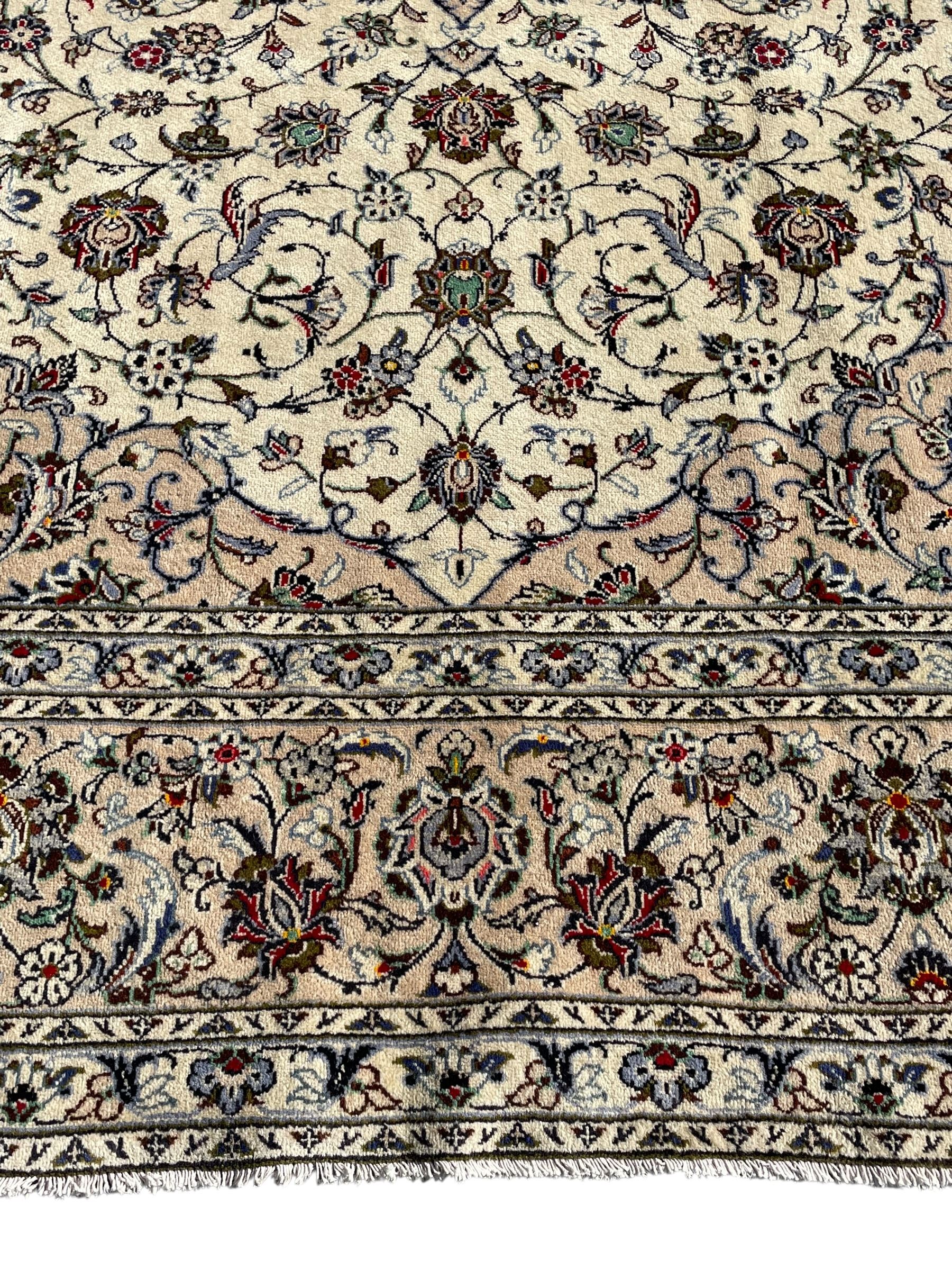 Persian Kashan ivory ground carpet, central rosette medallion with projecting palmettes, densely decorated field depicting trailing leafy branches and stylised plant motifs, the border with similar scrolling pattern, within floral pattern guard stripes 