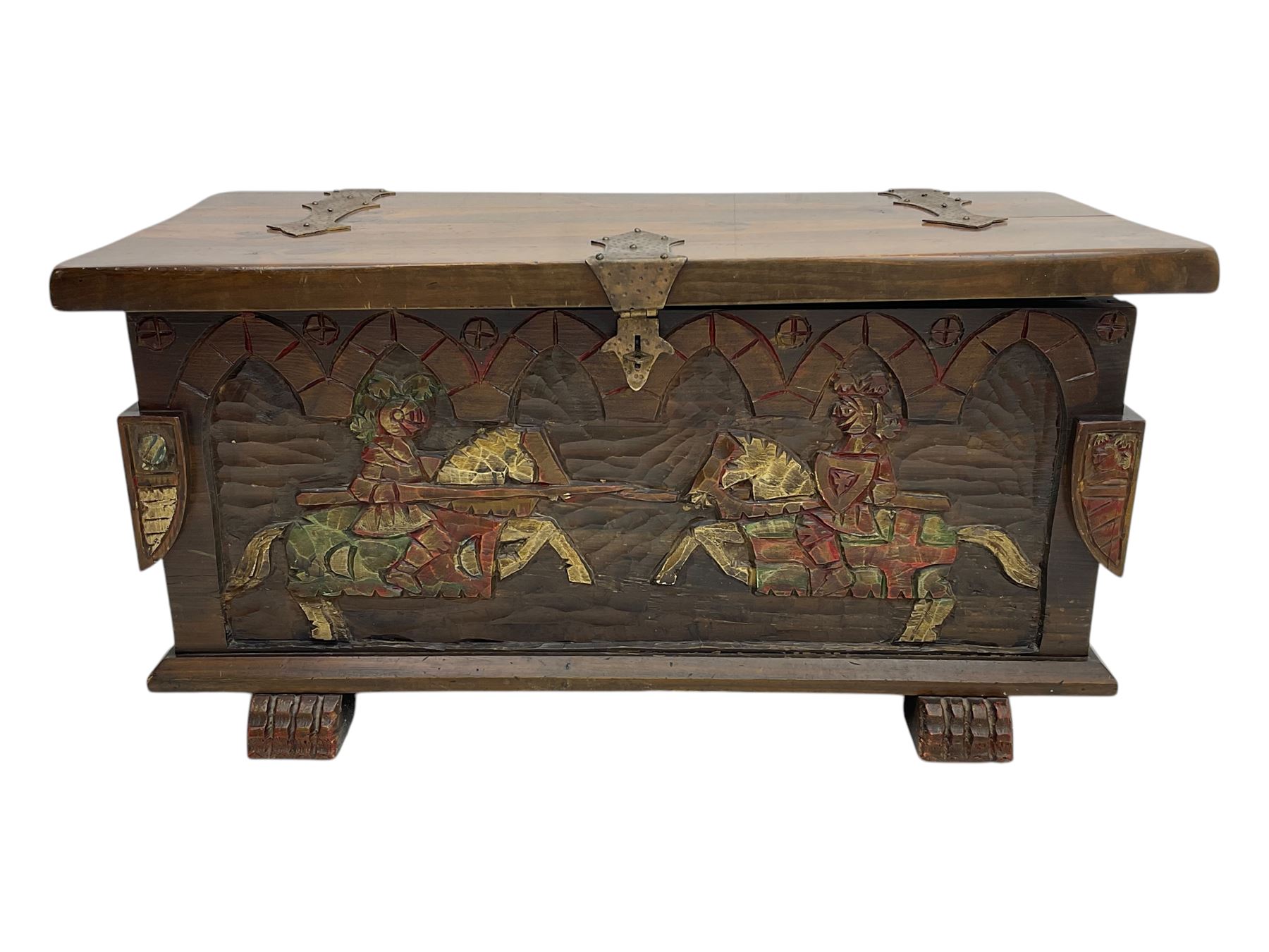 Mid-20th century carved hardwood chest, the rectangular hinged top with decorative strap hinges, the front and sides deeply carved with medieval-style jousting knights, heraldic shields, and architectural elements, raised on shaped block feet