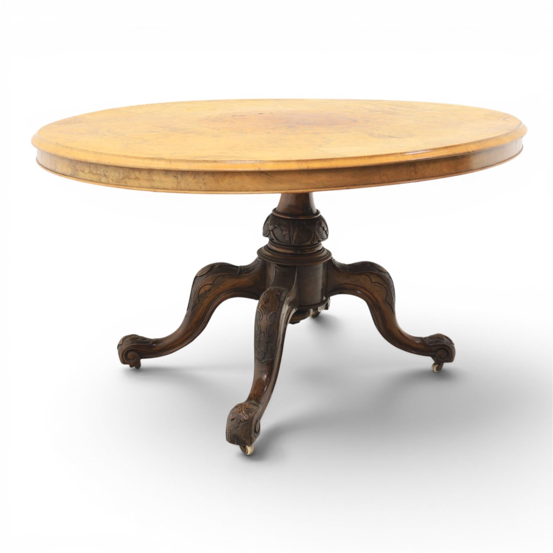 Victorian figured walnut oval loo tilt top table, raised on a heavily carved quatriform base with scrolled supports, terminating in castors