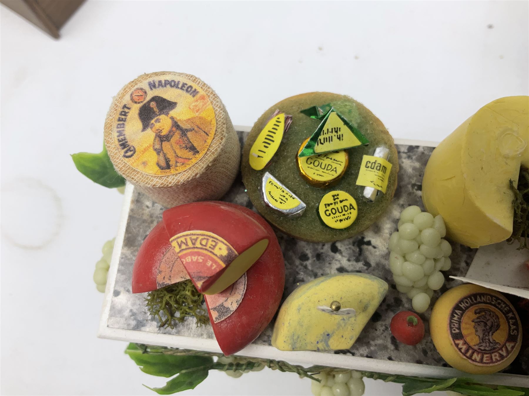 Collection of miniature dolls house grocery shop furniture and produce, comprising glazed corner counter with brass style cash register, shelved preserves unit, pies, cheese board and trays of meats, pair of white cheese counters, biscuit display with six tins, glazed preserves cabinet, pair of shop chairs and four further cheese boards complete with grapes and label flags