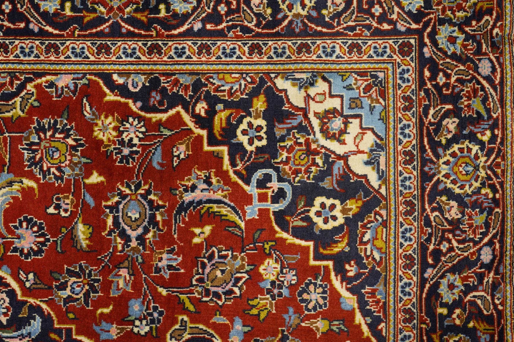 Central Persian Kashan hand-knotted rug, madder red field with central articulated cusped medallion in ivory and sky blue, issuing network of scrolling arabesques palmettes and floral sprays with conforming spandrels, indigo main border of scrolling vinery and stylized palmettes, framed by multiple guard borders, with cotton fringe ends  