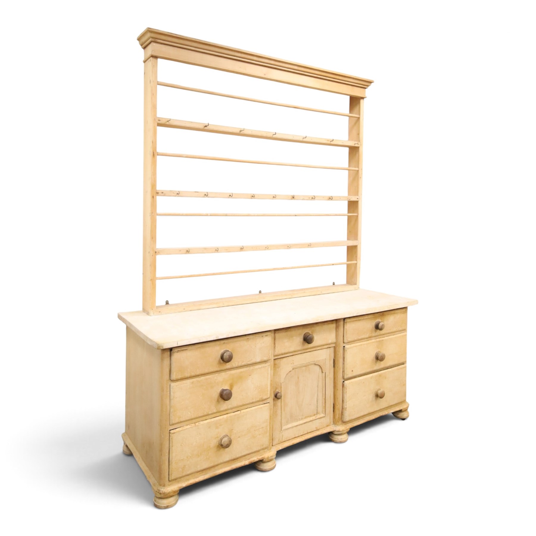 Victorian stripped pine dresser base with plate rack, the four-tier plate rack with supporting rails and hooks, the inverted-breakfront base fitted with seven assorted graduating drawers surrounding a recessed cupboard, on compressed bun feet with castors