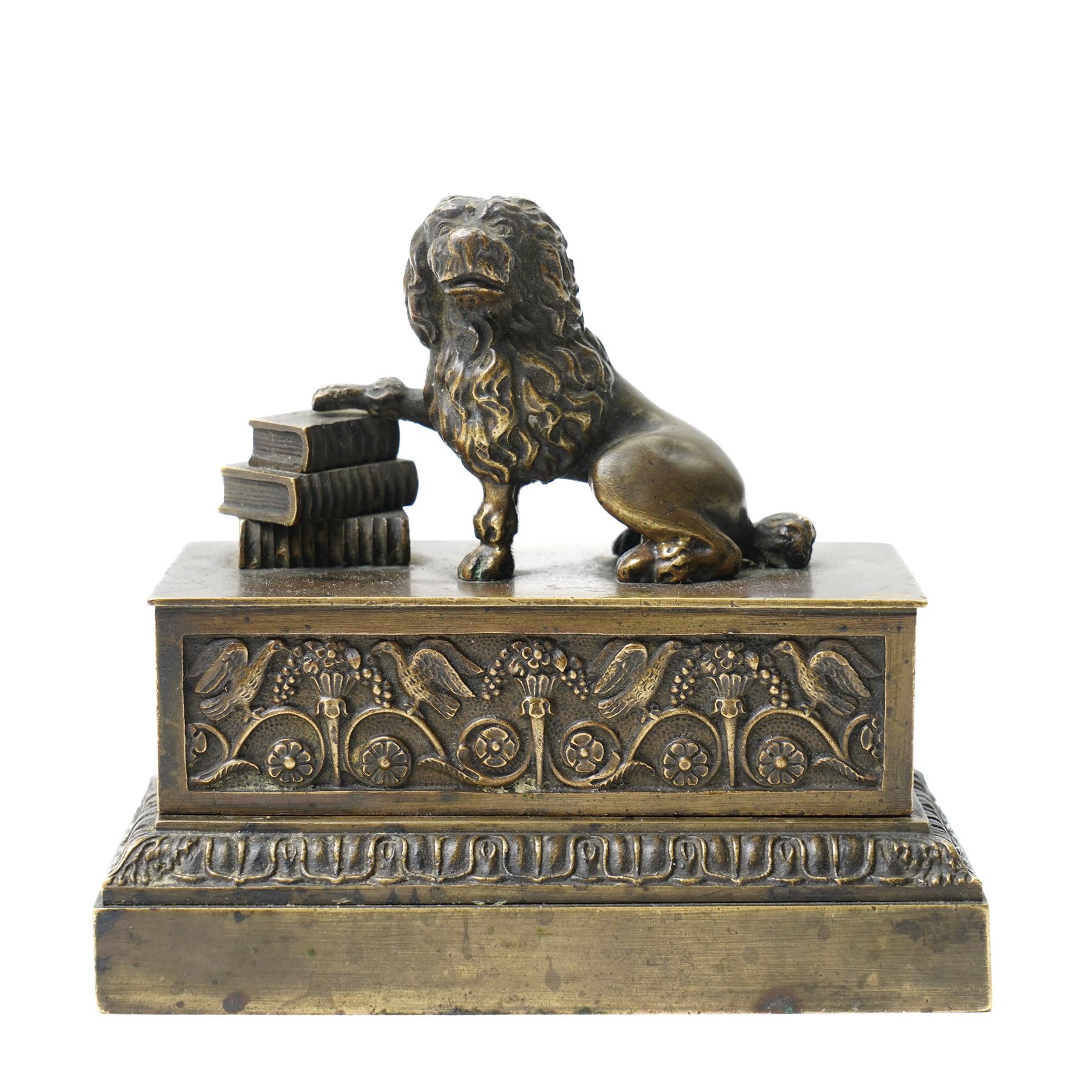 19th-century Grand Tour patinated bronze inkwell, surmounted by a recumbent lion, the beast modelled in the antique manner with one paw resting upon a stack of books, emblematic of strength, learning and guardianship. The stepped rectangular base cast with classical foliate friezes, fitted with multiple apertures and an inset ceramic inkwell and pounce pot; an erudite decorative object in the Grand Tour tradition, drawing on Renaissance and Roman prototypes, W12.5cm H11.5cm