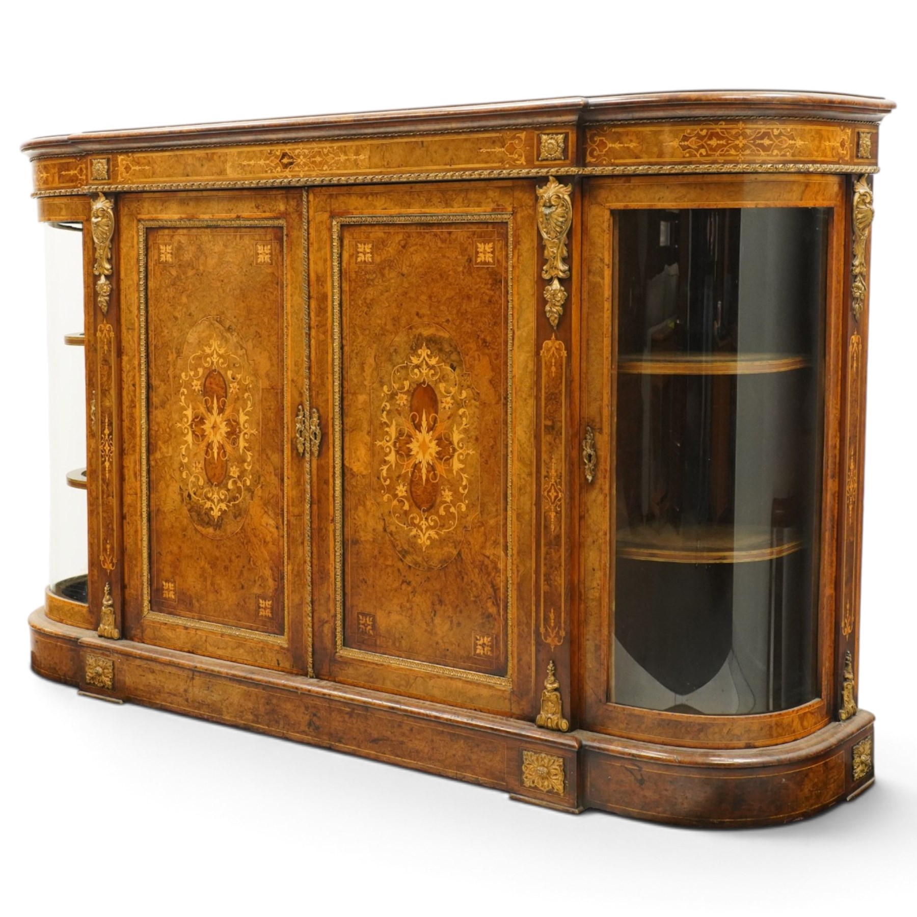 Large fine Victorian figured and burr walnut credenza, the breakfront D-shaped top with moulded edge, over an ornate frieze with inlaid Aesthetic Movement scrolling and geometric ornament, applied with gilt-metal ribbon-twist moulding and mounts, the central panelled doors profusely inlaid with scrolling foliate motifs and stringing, flanked by gilt-metal cartouche and scrolling acanthus corbel mounts, with flanking glazed bow-front side doors, on skirted base with further gilt metal foliate mounts