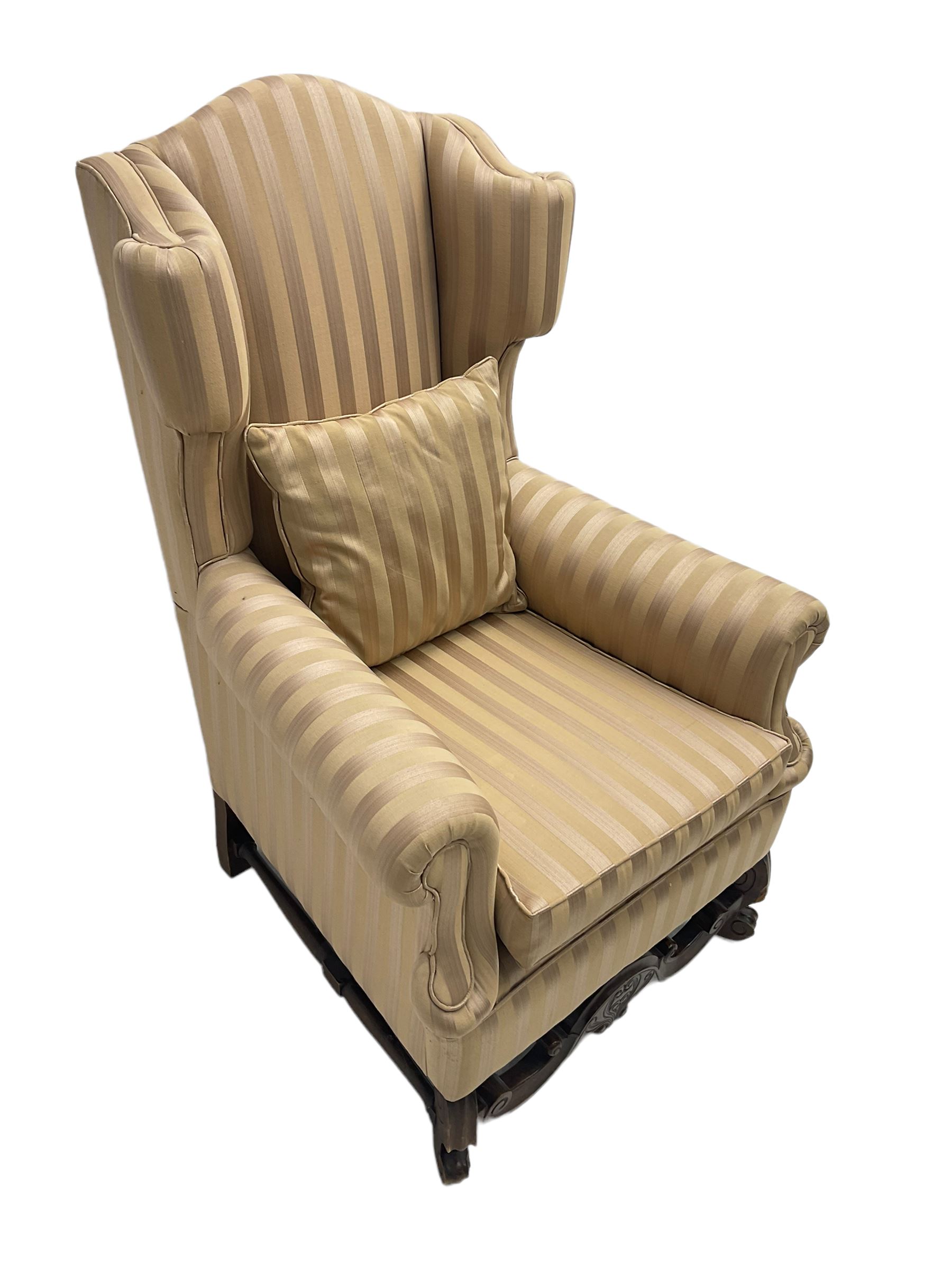 William and Mary style wingback armchair, upholstered in gold striped fabric, on walnut scroll carved supports joined by scroll and foliate carved middle rail, turned H-shaped stretchers, on castors