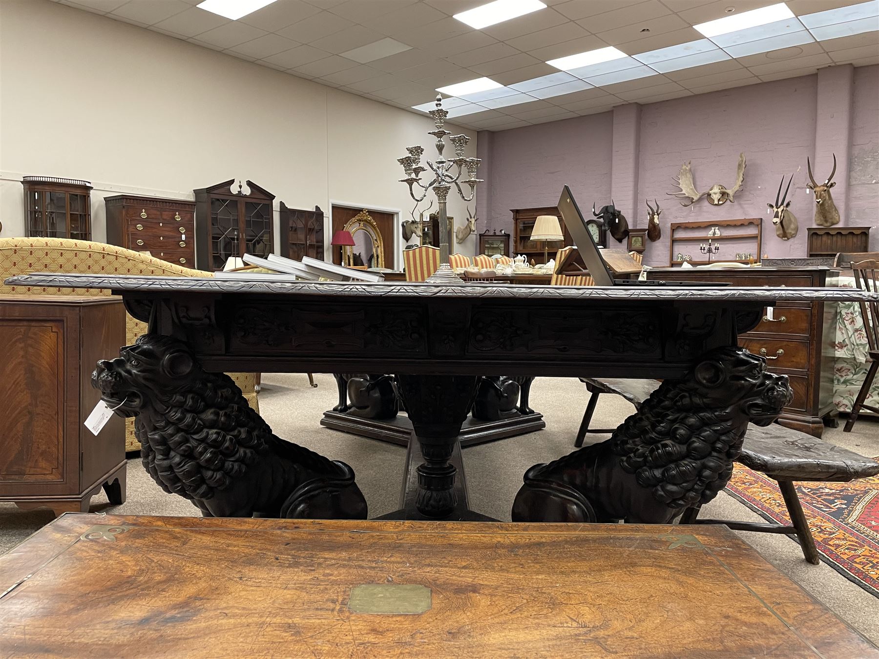 Large 19th century carved oak library table, the rectangular top with lunette carved edge over cartouche panelled and carved  scroll frieze, the central tapering rectangular support relief carved with masks and garlands, on four carved seated lion supports at each corner, on flat angular stretcher with recessed castors stamped ‘Bartholomew & Fletcher, London’