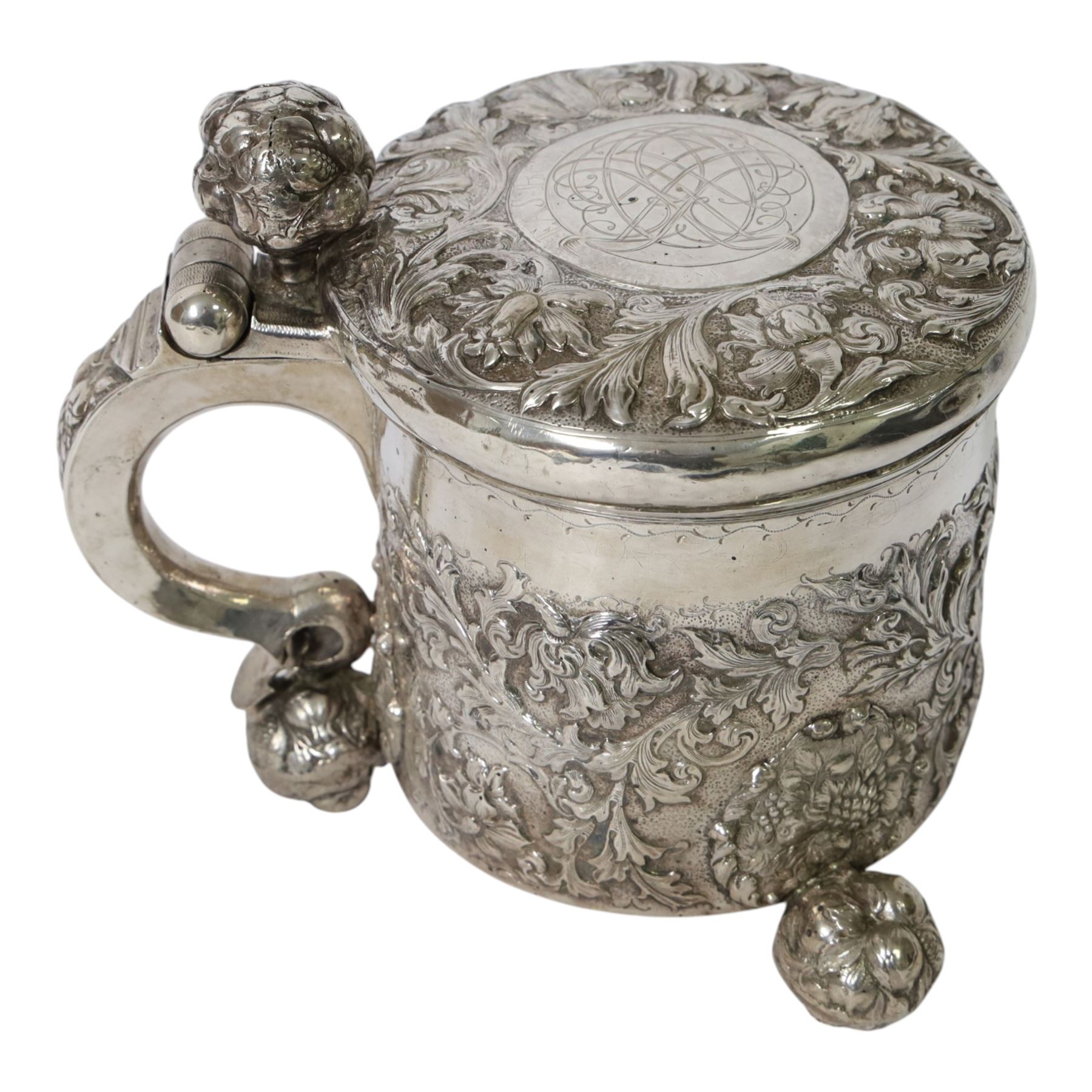 Northern European silver peg tankard, probably late 18th/early 19th century, of cylindrical form, the body repoussé decorated with scrolling and blossoming acanthus, fitted with scroll handle with shield terminal, the hinged cover with conforming repoussé decoration surrounding a central circular panel engraved with monogram beneath initials EASBGD, opening with ball thumbpiece detailed with fruits, raised upon three confirming ball feet mounted with shaped panels also detailed with fruits, bearing indistinct mark beneath, H19cm